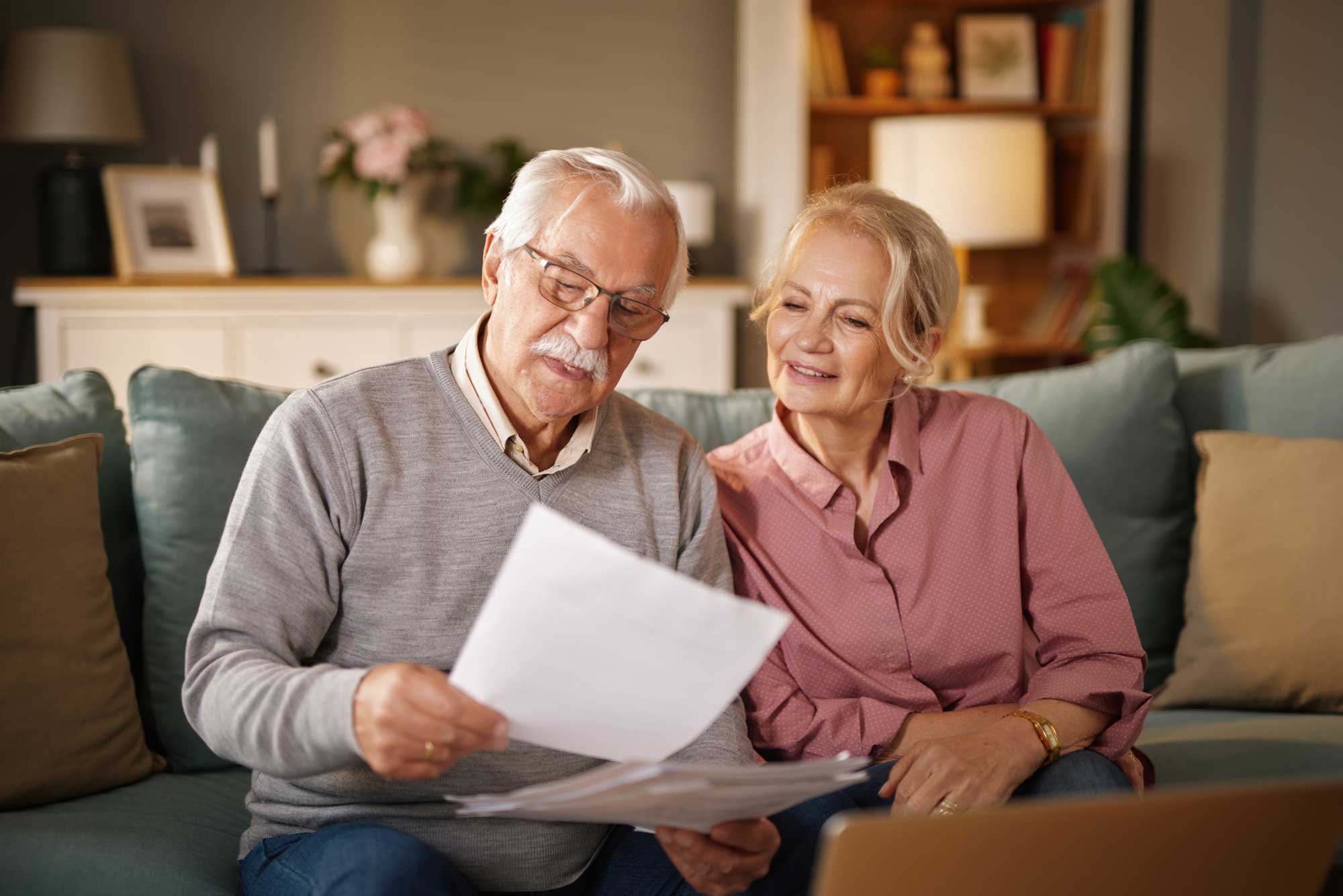 an-elderly-man-wearing-glasses-reads-paperwork-while-sitting-with-his-wife-on-a-sofa